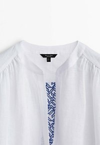 Blouse blanche texturée avec un col rond, des épaules plissées et une doublure intérieure bleue à motifs visible à l'ouverture du col.