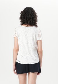 Haut en dentelle blanche avec motifs floraux, manches courtes et col rond, associé à une jupe noire plissée. Texture lisse, design décontracté.