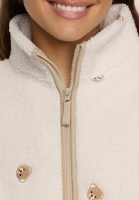 Nærbillede af en smilende person iført en cremet fleecejakke med en beige lynlås og små broderede teddybjørneansigtter.