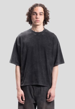 Jeune personne aux cheveux bouclés portant un t-shirt noir ample et délavé avec un pantalon assorti, debout devant un fond uni gris clair.