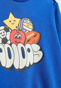 Sudadera azul con un diseño gráfico divertido que incluye pelotas deportivas al estilo de dibujos animados, una manzana y una estrella. Logotipo de "Adidas" en blanco en negrita.