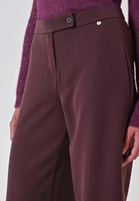 Pantaloni su misura bordeaux con frontale piatto, due tasche laterali e chiusura a un bottone. Tessuto liscio con una leggera lucentezza.