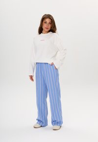 Weißes langärmliges Hemd mit rosa Text, kombiniert mit hellblauen weiten Hosen mit weißen und rosa vertikalen Streifen. Die Schuhe sind hellbeige.
