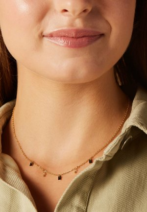 Collier en chaîne dorée comportant de petits charms rectangulaires et ronds. La chaîne est délicate, avec une texture lisse, portée près de la clavicule.