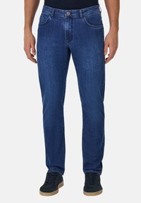 Jeans de mezclilla azul con un diseño de pierna recta, cinco bolsillos, acentos de costura y un cierre de botón. Llevados con zapatillas negras.