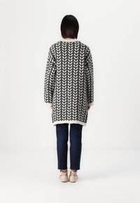 Cardigan a maglia in bianco e nero, con un motivo a zigzag verticale, maniche lunghe e vestibilità comoda, abbinato a jeans scuri e scarpe chiare.