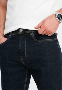Pantalones vaqueros de mezclilla azul oscura con costuras amarillas, diseño de cinco bolsillos y un botón de metal en la cintura, presentando un primer plano de la textura de la tela.