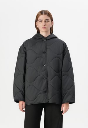 Een model in een zwart gewatteerd jasje met capuchon staat tegen een effen witte achtergrond, met een eenvoudige knoopsluiting.