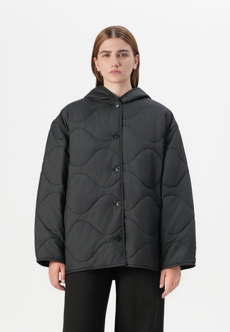 Ein Modell trägt eine schwarze, gesteppten Jacke mit Kapuze und steht vor einem schlichten weißen Hintergrund, die einen einfachen Knopfverschluss aufweist.