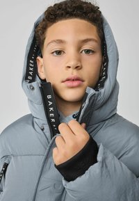 Veste doudoune grise avec un extérieur texturé, une capuche surdimensionnée et des emblèmes "BAKER" noirs sur les zips et les poignets. Coupe ajustée avec des accents élastiques.