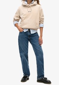Felpa in corduroy beige con tasca frontale e coulisse, indossata sopra una camicia azzurra a maniche lunghe, abbinata a jeans blu a gamba dritta. Scarpe nere.