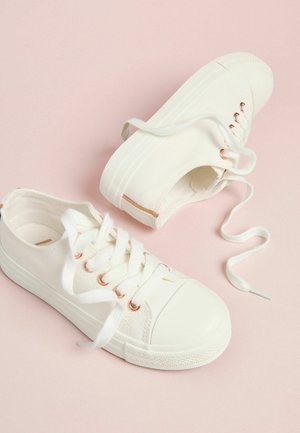 Balti auduma sporta apavi ar apaļu purngalu, gumijas zolīti un rozā zelta skabām. Šņores austi caur skabām, atrodas uz rozā fona.