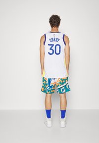 Giovane in piedi di spalle che indossa una maglia da basket bianca "Curry 30" e pantaloni corti a stampa colorata di delfini, calzini blu e scarpe bianche.