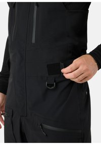 Zwarte geïsoleerde overall met een ritssluiting pocket, verstelbare bandjes, klittenbandsluiting en stevige stof met een gladde textuur.