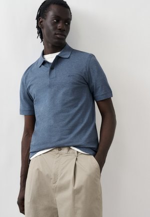 Fiatal, sötét bőrű férfi dreadlockkal, kék rövid ujjú galléros pólót és bézs nadrágot visel, oldalra néz, kezét a zsebében tartva.