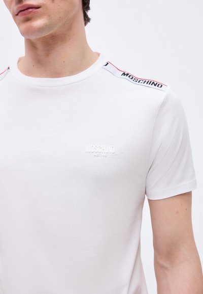 Νεαρός άνδρας που φοράει λευκό t-shirt Moschino Active με λογότυπο στο στήθος και διακοσμητική ταινία με το εμπορικό σήμα στους ώμους.
