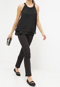 Czarny bezrękawnik z marszczonym dekoltem, warstwowa konstrukcja; w połączeniu z dopasowanymi czarnymi spodniami i płaskimi czarnymi butami, trzymająca kopertówkę.