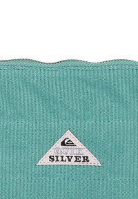 Turquoise stof met ribbelstructuur, voorzien van een witte driehoekige logo patch met daarop in vetgedrukte zwarte letters "QUIKSILVER" in het midden.