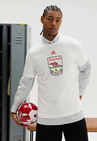 Wit T-shirt met korte mouwen met het Arsenal-logo en rode accenten, gedragen over een gestreept shirt met lange mouwen. Houdt een rood-witte voetbal vast.