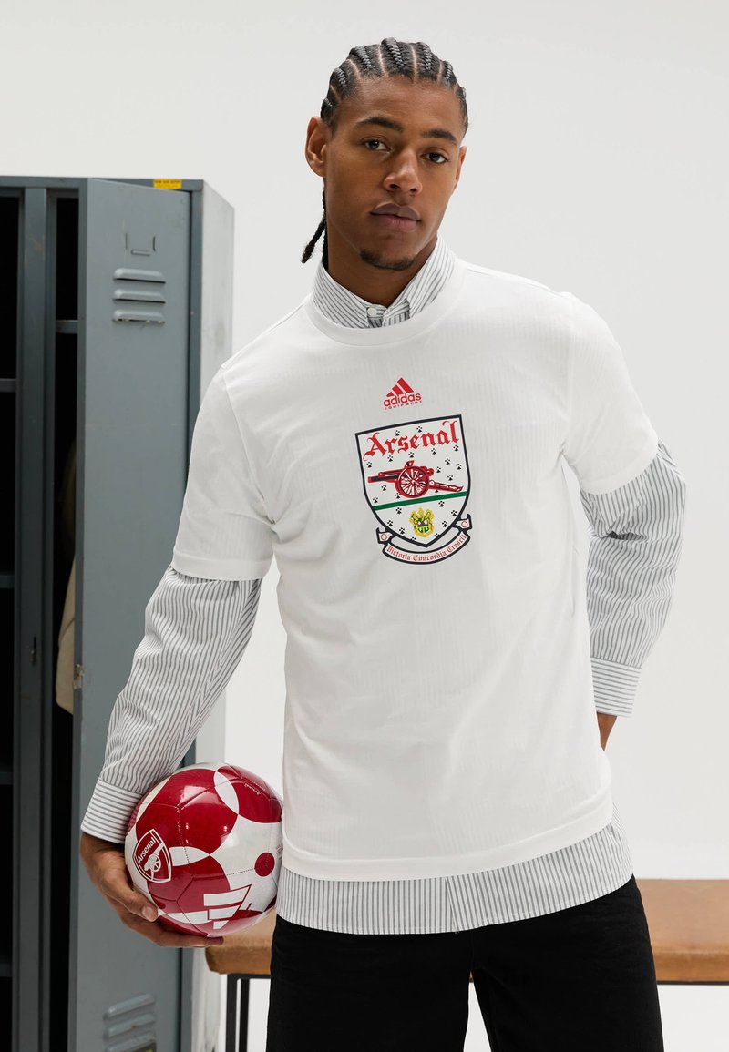 Wit T-shirt met korte mouwen met het Arsenal-logo en rode accenten, gedragen over een gestreept shirt met lange mouwen. Houdt een rood-witte voetbal vast.