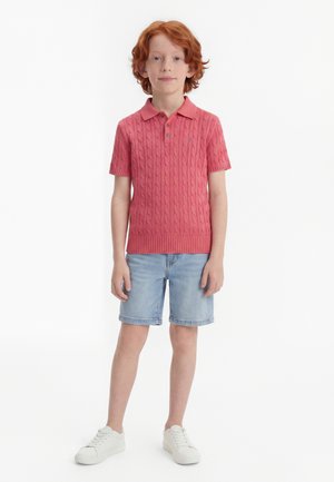 Rude dziecko stoi, ubrane w koralowe, krótki rękaw, dzianinowe polo, jasnoniebieskie szorty jeansowe oraz białe trampki na białym tle.