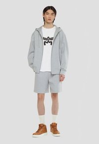 Sweat à capuche gris zippé, t-shirt blanc avec un motif noir, short gris, et baskets montantes marron à semelles blanches. Tissu doux et décontracté.