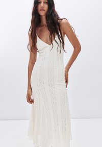Abito bianco in pizzo con design a V, busto aderente, trama semi-trasparente e gonna fluente con motivi verticali.