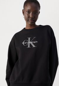 Čierna mikina so šedým logom "CK" a textom "Calvin Klein". Vyrobená z jemnej látky s voľným strihom a okrúhlym výstrihom.