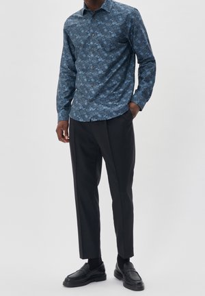 Mann trägt blaues langärmliges Hemd mit Blumenmuster, schwarze maßgeschneiderte Hose, schwarze Socken und schwarze Lederloafer, steht mit einer Hand in der Tasche.