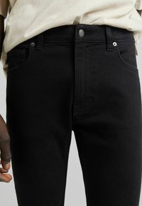 Jeans de mezclilla negra con un ajuste slim, cintura alta, cierre de botones, herrajes metálicos y bolsillos frontales. La textura es suave y sólida.