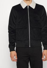 Svart kordjacka med vit fuskpälskrage, framzip med dragkedja och två nedre påsydda fickor. Ribbad ärmslut och nederkant.