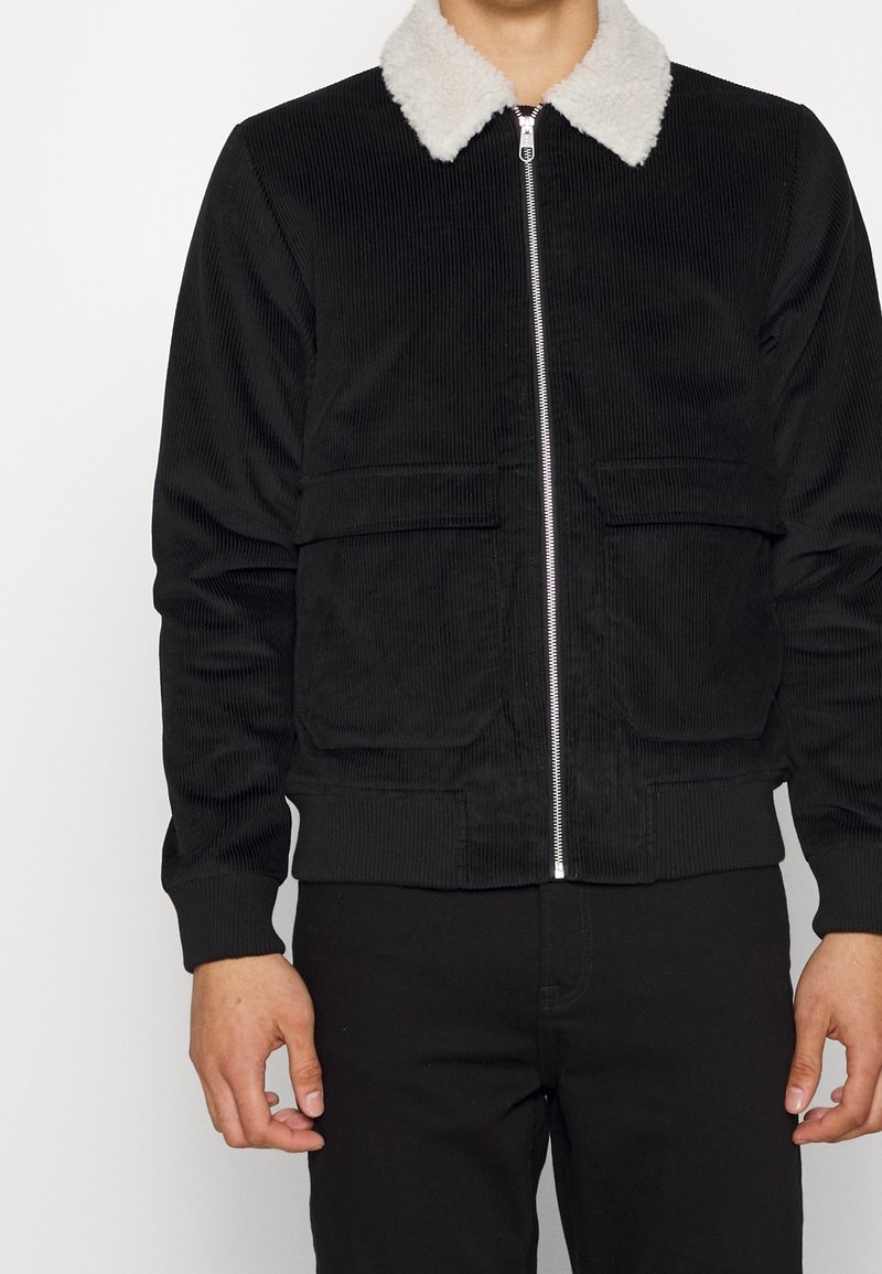 Svart kordjacka med vit fuskpälskrage, framzip med dragkedja och två nedre påsydda fickor. Ribbad ärmslut och nederkant.