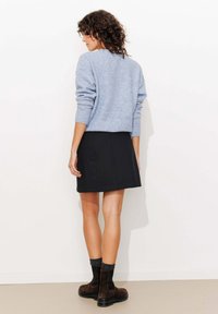 Femme aux cheveux bouclés portant un pull bleu clair, une minijupe noire, des chaussettes grises et des bottines brunes, debout contre un mur blanc.
