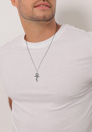 Silberne Halskette mit einem Ankh-Anhänger, getragen über einem weißen, eng anliegenden T-Shirt. Der Anhänger zeigt filigrane Details an einer dünnen Kette.