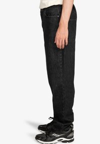 Černé džínové kalhoty s rovnými nohavicemi, pětikapsovým střihem a jemnou texturou. Nošeny s černými sportovními botami.