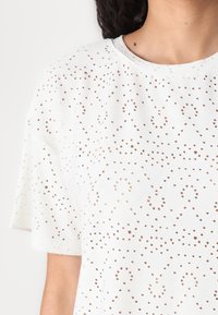 Haut blanc à manches courtes avec un col rond, présentant un motif complexe de petites découpes et un tissu doux et texturé.