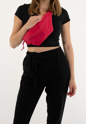 Punainen kangasvyölaukku, joka on käytössä ristiin, varustettuna vetoketjulla, kohotetulla logolla ja tyylikkäällä, kulmikkaalla muotoilulla. Mustat housut, joissa on nauhaompelu.