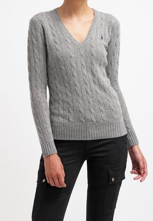 Personne portant un pull gris ajusté en maille torsadée avec col en V et un pantalon noir avec poches cargo à boutons, visible du cou jusqu'aux hanches.