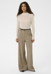 Sieviete ar gariem cirtainiem matiem, valkājot caurspīdīgu baltu rakstainu garroku kreklu, kas iestiprināts augsta jostas plaša piegriezuma bēšās biksēs ar melnu jostu.