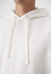 Balta kapucīna klozīte, kas izgatavota no maiga auduma ar klasisku dizainu. Aprīkota ar regulējamu kapuci un mazu, izšūtu Nike logo.