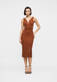 Γυναίκα που φοράει φορεμα midi χωρίς μανίκια σε σκουριά χρώμα με βαθύ V ντεκολτέ και σκίσιμο μπροστά, συνδυασμένο με μαύρα πέδιλα με λουράκι στον αστράγαλο, στέκεται μπροστά σε λευκό φόντο.