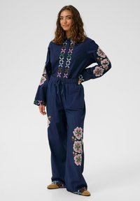 Femeie stând în picioare cu mâna pe șold, îmbrăcată într-un salopetă albastru închis cu broderie florală colorată pe mâneci, parte frontală și picioare, asortată cu pantofi bej.