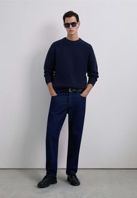 Suéter de punto azul marino, jeans oscuros de corte holgado, zapatos negros y gafas de sol. La modelo está de pie contra un fondo liso con las manos en los bolsillos.