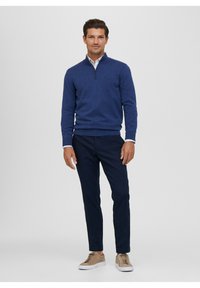 Maglione a maglia blu navy con colletto con zip, abbinato a pantaloni scuri e sneakers color marrone chiaro. L'ambientazione è neutra.