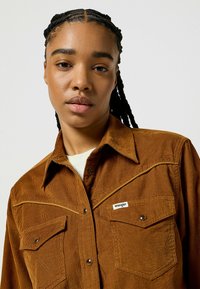 Jeune femme avec des cheveux tressés portant une chemise en velours côtelé marron de Wrangler avec des poches avant et des boutons pression en métal sur un fond uni.
