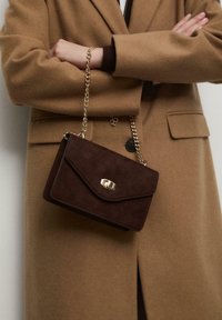 Brązowa skórzana torebka z zapięciem na złotą klamrę, z klapą i łańcuchowym paskiem. Torebka jest trzymana obok beżowego płaszcza z detalem w postaci guzików.