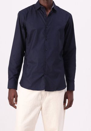 Camisa azul marino abotonada con cuello clásico, mangas largas y un sutil brillo, combinada con pantalones de color claro. Diseño sencillo sin estampados.