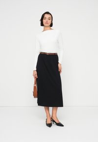 Μαύρη μακριά φούστα με ζώνη σε ταμπά απόχρωση, συνδυασμένη με λευκό μακρυμάνικο τοπ. Το μοντέλο φοράει μαύρες γόβες μυτερές και κρατά καφέ τσάντα.