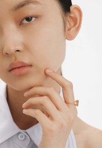 Anillo de gema triangular con banda de oro rosa. La mano del modelo descansa sobre la barbilla, mostrando una piel suave y una sencilla camisa de cuello.