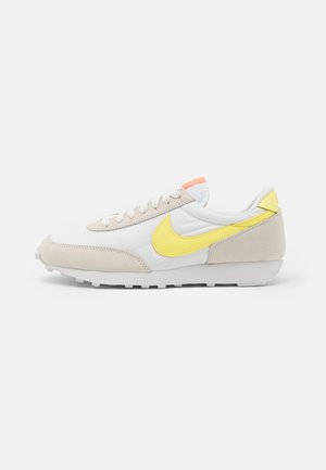 Biały i beżowy sneaker z żółtym logo Nike, pomarańczowym akcentem na języku i białą gumową podeszwą, pokazany z profilu na białym tle.
